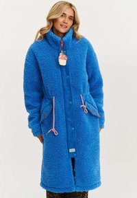 Modrá fleecová bunda s vysokým golierom, patentnými gombíkmi a zamatovými nášivkami, s šnúrkou v páse a dekoratívnou visačkou. Dve predné vrecká.