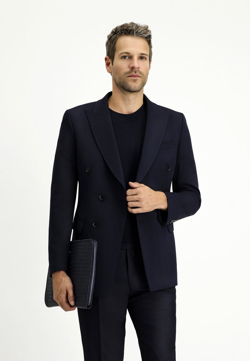 Blazer azul marino de corte cruzado con solapas en pico, combinado con una camisa negra y pantalones a juego. Sosteniendo una funda de tableta negra texturizada.