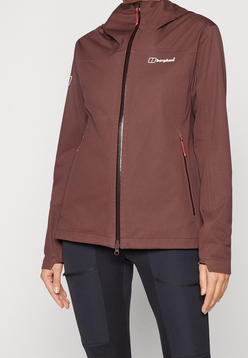 Femme portant une veste Berghaus marron à fermeture éclair avec des poches latérales zippées et un pantalon ajusté foncé avec des poches cuisses zippées.