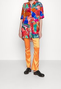 Person som bär en färgglad djurmönstrad skjorta, blanka orange byxor och svarta nitarklädda träskor, stående mot en enfärgad bakgrund.