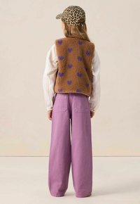 Gilet en polaire marron avec des motifs de cœurs violets, chemise à manches longues blanche, et pantalon lavande ample, associés à une casquette à imprimé léopard.