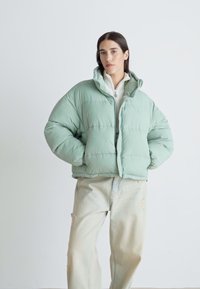Giacca imbottita verde chiaro con colletto alto, chiusura con zip e pannelli cuciti, abbinata a pantaloni in denim chiaro e consumato.
