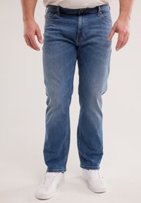 Férfi világoskék egyenes szárú farmernadrágban, fehér tornacipőben és begombolt bézs ingben, karjaival lazán oldalt állva.