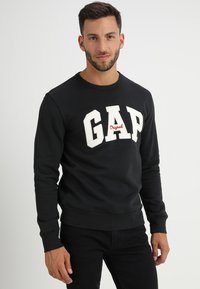 Black sweatshirt made of cotton featuring large white "GAP" logo and "Original" in red. It has a crew neckline and ribbed cuffs.