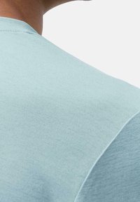 Närbild av en persons axel och nacke som bär en ljusblå t-shirt med rund halsringning mot en enfärgad vit bakgrund.