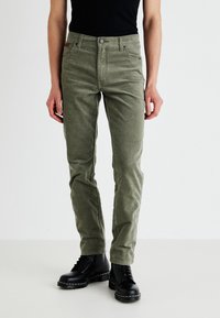 Olíva zöld kordbársony nadrág, slim fit fazonban, elülső zsebekkel, cipzáros főrésszel és ezüst gomb részletekkel. Fekete fűzős csizmával viselve.