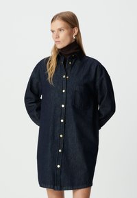 Kobieta w ciemnoniebieskiej długiej sukience koszulowej z z�łotymi guzikami i brązowym golfem, stojąca na tle białego tła.