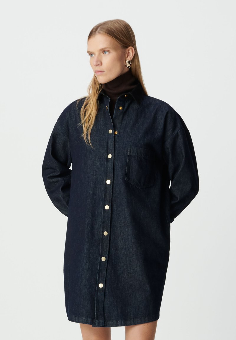 Kobieta w ciemnoniebieskiej długiej sukience koszulowej z złotymi guzikami i brązowym golfem, stojąca na tle białego tła.