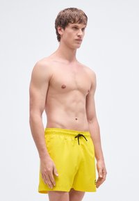 Dzelteni peldbikses ar elastīgu vidukļa joslu, melnu auklu un smalku logotipa detaļu. Gluda tekstūra, dizains virs ceļgala.