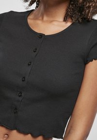 Mujer que lleva un top corto negro de manga corta acanalado, con seis botones negros en frente.