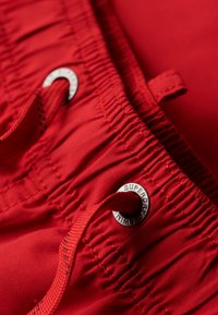 Shorts de bain rouges avec taille à cordon de serrage, texture froncée, et œillets métalliques avec logo en relief. Tissu lisse et léger.