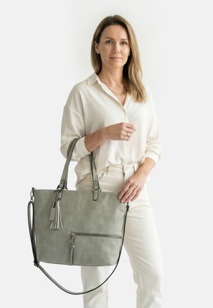 Woman with light brown hair wears white blouse and pants, holding large gray handbag with tassel and zipper pocket against plain background.
