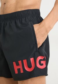 Juodi maudymosi šortai pasižymi lengvu audiniu ir elastinga juosmeniu. Šoninė kišenė ir ryškus raudonas užrašas "HUG" yra išskirtiniai bruožai.