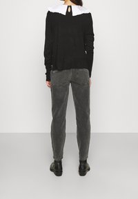 Musta pitkähihaiseksi paita, jossa on valkoinen röyhelökaulus ja takana napitus, yhdistettynä harmaisiin korkeavyötäröisiin housuihin ja mustiin saappaisiin.