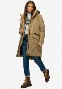 Beige gevoerde parka met een capuchon, ritssluiting en twee voorzakken. Draagt over een gebreid patroontrui, gecombineerd met opgerolde spijkerbroek in het blauw en bruine laarzen.