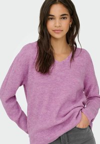 Jeune femme aux longs cheveux foncés portant un pull côtelé lilas et un jean gris, posant avec une main dans la poche devant un fond blanc.