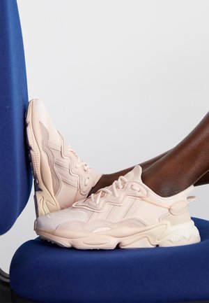 Person iført lyserøde atletiksneakers, hviler fødderne på en blå stol og læner sig mod en hvid baggrund.