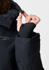 Hand adjusting black jacket hood with drawstring toggle, wearing a thumb hole glove under the sleeve.