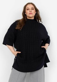 Marinblå kabelstickad oversized tröja med korta ärmar och hög krage, med en ribbad nederkant. Matchad med grå avsmalnande byxor.