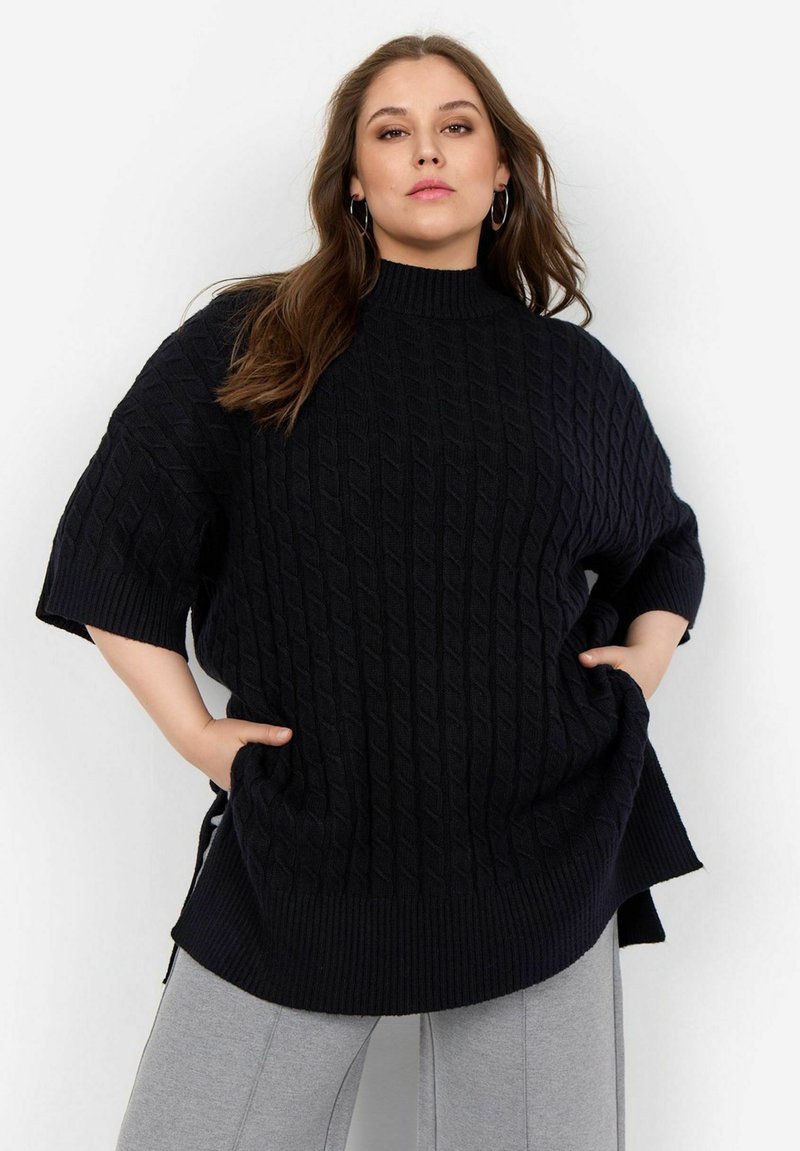 Marinblå kabelstickad oversized tröja med korta ärmar och hög krage, med en ribbad nederkant. Matchad med grå avsmalnande byxor.