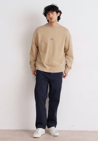 Felpa beige con collo rotondo e polsini a coste, caratterizzata da un piccolo logo sul petto. Abbinata a pantaloni blu navy e sneakers bianche.