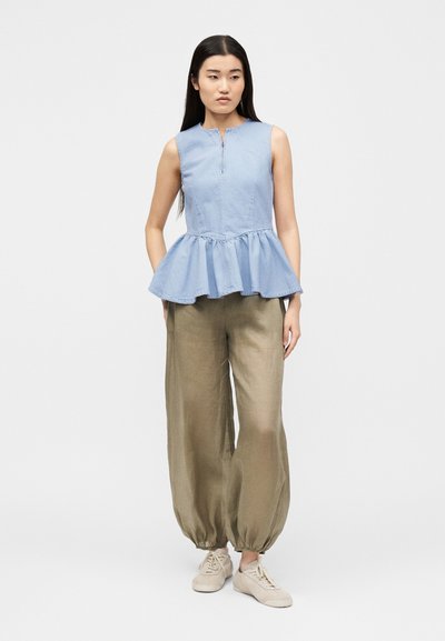 Femme aux longs cheveux noirs portant un top péplum bleu clair sans manches, un pantalon large marron et des baskets beiges, debout avec les mains dans les poches.