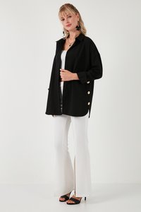 Ženska, oblečena v ohlapno črno srajco z zlatimi gumbi na strani, bele hlače z sprednjimi razporki, črne sandale s peto.