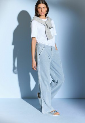 Valkoinen T-paita, harmaa neule solmittuna olkapäiden ympärille, siniraitaiset leveät housut ja hopeiset sandaalit, vaaleansinistä taustaa vasten.