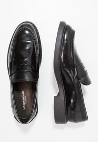 Mocassins de couro preto com um acabamento brilhante, apresentando uma faixa decorativa e costura em contraste. Solas de borracha planas e design de bico arredondado.