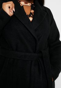 Abrigo de lana negro con solapa ancha, cintura ceñida con cinturón y bolsillos laterales. Blusa estampada visible por debajo con un detalle de ojal en el escote.