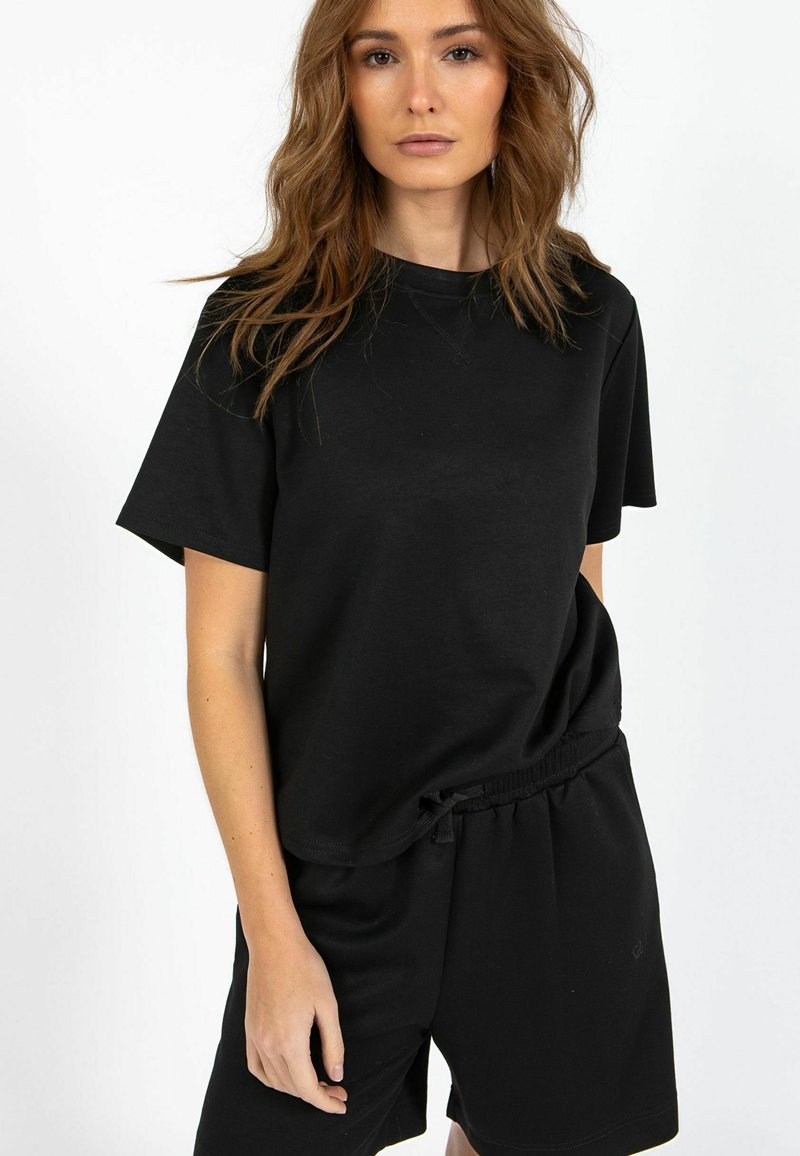 Woman with wavy brown hair wearing a plain black short-sleeve shirt and black shorts against a white background.