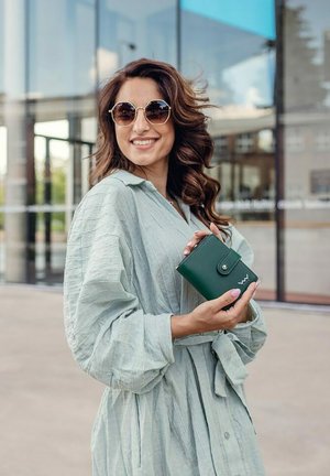 Green wallet held in a hand, made of leather with a button closure, featuring a subtle logo. The model wears a light green textured shirt.