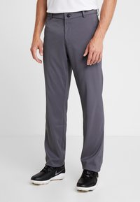 Man i grå skräddarsydda byxor och svarta sportskor står mot en vit bakgrund, med händerna avslappnat vid sidorna.