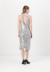 Vestido prateado com lantejoulas, comprimento até o joelho, design sem mangas e um profundo decote em forma de buraco-chave nas costas. Sapatos de salto altos pretos complementam o visual.