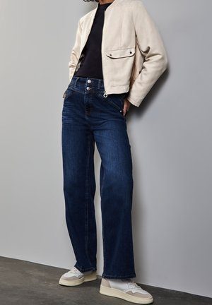 Person iført en beige jakke, sort top, mørkeblå vidde jeans og hvide sneakers, står op ad en grå væg.