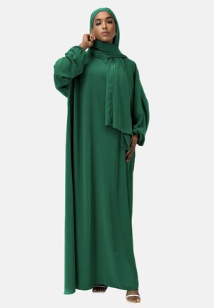 Un hijab vert et une longue robe fluide en tissu léger, avec des manches bouffantes et une écharpe nouée au cou.