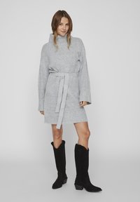Robe pull grise texturée avec des manches longues et une taille ceinturée. Associée à des bottes noires en daim jusqu'aux genoux, avec un talon carré.