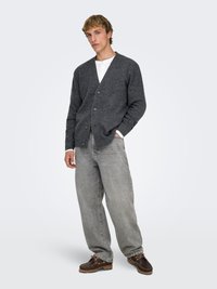 Gray wool cardigan with a V-neck and button closure, worn over a white henley. Paired with loose light gray jeans and brown shoes.