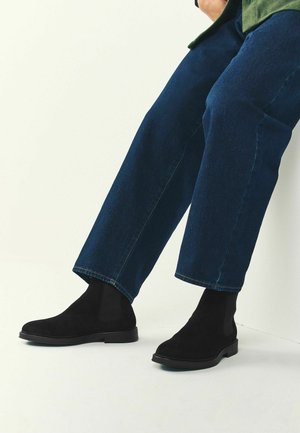 Juodos zomšos kulkšninių botai su elastinėmis šoninėmis dalimis ir plokščiu padu, derinami su trumpomis mėlynomis džinsinėmis kelnėmis. Minkšta tekstūra ir struktūrinė forma.