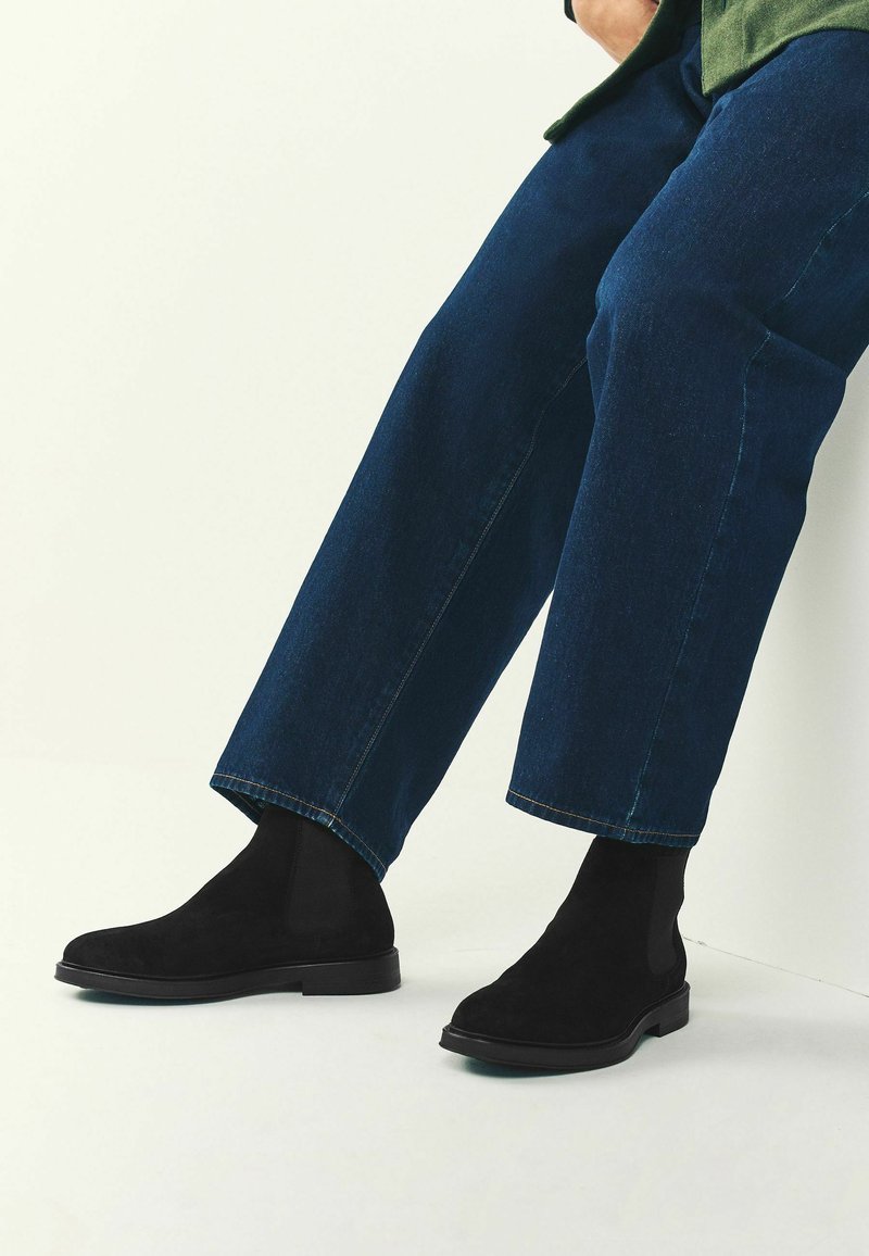 Mustad sametist nahast pahkluusaapad elastsete külgpaneelidega ja lamedate talladega, kombineeritud lühikeste siniste denimpükstega. Pehme tekstuur ja struktureeritud kuju.