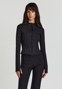 Donna che indossa una giacca nera aderente con zip e leggings abbinati con un sottile motivo di leopardo scuro, in piedi di fronte a uno sfondo chiaro e uniforme.