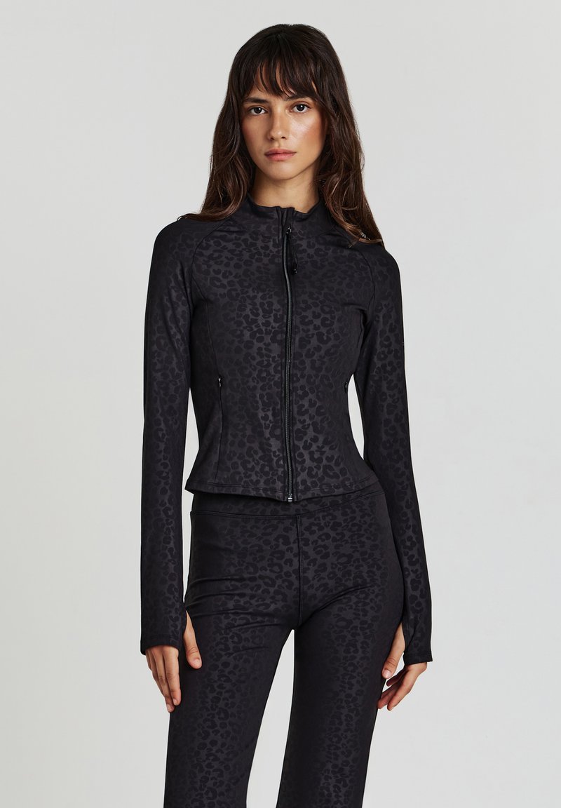 Donna che indossa una giacca nera aderente con zip e leggings abbinati con un sottile motivo di leopardo scuro, in piedi di fronte a uno sfondo chiaro e uniforme.