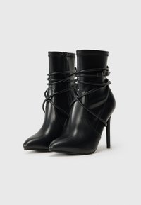 Pár čiernych kožených čižiem s vysokým opätkom, s špicatými špičkami, obtočené tenkými pásikmi a malými postrannými prackami, stojace na bielom pozadí.