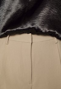 Gros plan de la taille d’un pantalon beige avec des passants de ceinture sous un haut en tissu noir brillant.
