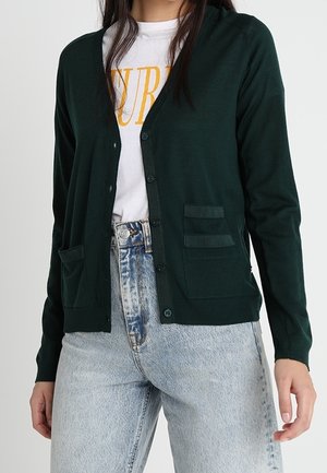 Femme portant un cardigan boutonné vert foncé sur un t-shirt blanc à motif, associé à un jean taille haute bleu clair.
