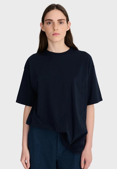 Jeune femme aux longs cheveux bruns portant un t-shirt ample bleu marine foncé et un pantalon assorti, debout devant un fond clair uni.