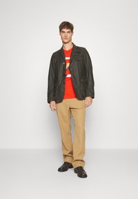 Dark green lightweight jacket with pockets, layered over a red graphic t-shirt. Beige trousers and brown shoes complete the look.