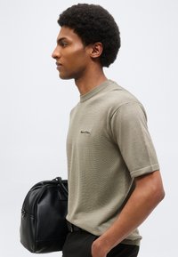 Άνδρας με κόμμωση αφρο, φοράει μπεζ πουκάμισο με κοντά μανίκια, κρατάει μαύρη δερμάτινη τσάντα, στέκεται σε προφίλ μπροστά σε απλό φόντο.