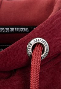 Weinroter Sweatshirt aus strukturiertem Stoff, mit einem silbernen Ösen und einem geflochtenen Kordelzug. Schwarzes Etikett mit weißem Text sichtbar.