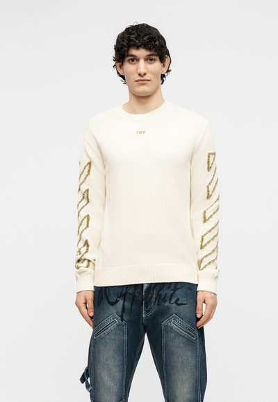 Άνδρας με σγουρά σκούρα μαλλιά, φοράει λευκό πουλόβερ με χρυσές διαγώνιες ρίγες στα μανίκια και μπλε τζιν παντελόνι με μαύρα σχέδια γραφής.